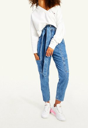 Nainen pukeutunut valkoiseen olkapäät paljastavaan pitkähihaiseen paitaan, korkeavyötäröisiin sinisiin farkkuihin vyöllä ja valkoisiin lenkkareihin, joiden pohjat ovat vaaleanpunaiset.