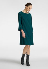 Türkisfarbnes Shiftkleid mit Dreiviertelärmeln, Trompetenärmeln und Knopfdetails. Kombiniert mit schwarzen kniehohen Stiefeln und minimalistischem Schmuck.