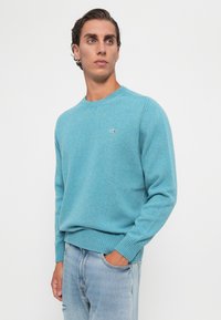 Junger Mann in einem türkisfarbenen Rundhalsausschnittpullover und hellblauen Jeans, mit einer Hand in der Tasche, schaut leicht zur Seite vor einem weißen Hintergrund.