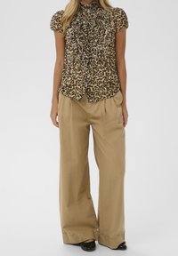 Blouse à imprimé léopard avec devant volanté, manches courtes et col montant, associée à un pantalon beige à jambes larges et des chaussures noires.