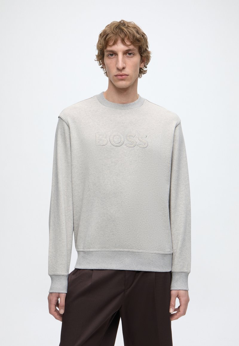 Jeune homme aux cheveux bouclés portant un sweat-shirt gris clair avec "BOSS" en relief sur la poitrine et un pantalon marron foncé, debout sur fond blanc.