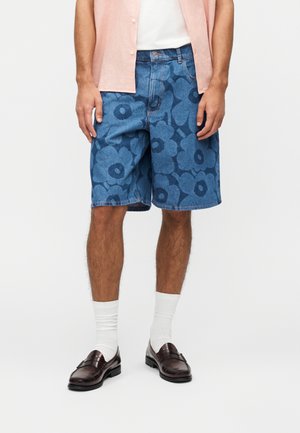 Mężczyzna ubrany w niebieskie krótkie spodenki dżinsowe z dużymi ciemnoniebieskimi kwiatowymi wzorami, białe skarpetki, brązowe skórzane mokasyny, białą koszulę oraz jasnoróżową otwartą koszulę.