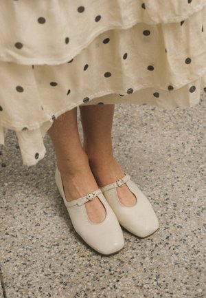 Pieds portant des chaussures Mary Jane couleur crème avec boucles sous une robe crème à pois noirs, debout sur un sol en terrazzo.