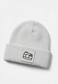 White ribbed knit beanie featuring a black camera graphic on a square patch. The texture is smooth with a folded cuff.