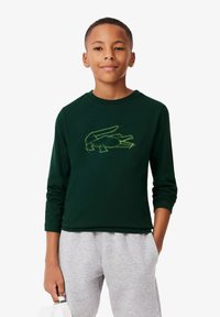 Felpa verde a maniche lunghe con collo rotondo e un logo geometrico giallo neon sul davanti, abbinata a pantaloni della tuta grigi.