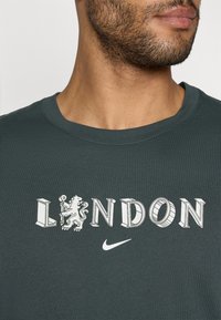 Tumši zaļš T-krekls ar burtu "LONDON" treknā baltā krāsā, uz kura ir lauvu grafika un mazs Nike logo zem tā, izgatavots no mīkstas kokvilnas auduma.