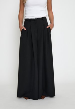Donna che indossa pantaloni neri a gamba larga con pieghe, in piedi con le mani nelle tasche, abbinati a un top bianco senza maniche e scarpe con fantasia.