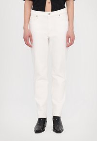 Weiße Denim-Jeans mit geradem Schnitt, mit Reißverschluss, fünf Taschen und dezenten Nahtdetails. Tragen mit schwarzem Schuhwerk.