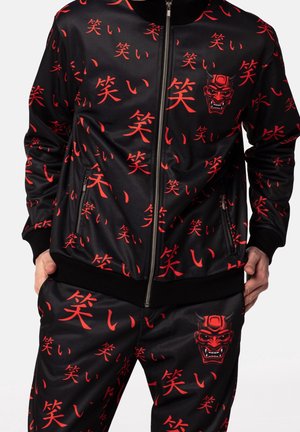 Veste de sport noire et pantalon avec des caractères kanji japonais rouges et des motifs de masque de démon rouge, fermeture éclair à l’avant et poches zippées sur la veste.