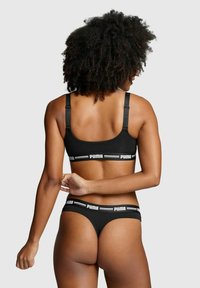 Schwarzes Dessous-Set mit einem Bralette mit verstellbaren Trägern und einem passenden Tanga. Beide Teile zeigen ein weißes Logo und gestreifte Details.