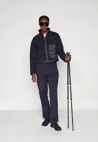 Temno modra skrajšana jakna iz teksturiranega flisa s sijočim detajlom na žepu, v kombinaciji z ujemajočimi temnimi hlačami in črno obutvijo. V roki drži pohodne palice.