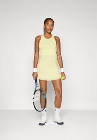 Vestido de ténis amarelo com um design de costas em raia, apresentando um corpete texturizado e uma renda na bainha. Pulsatórias brancas, sapatilhas e uma raquete de ténis.