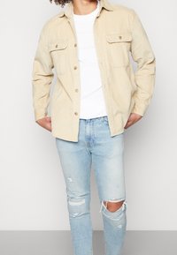 Beige kordflöjtskjorta med knappar och två bröstfickor, ihop med slitna ljusblå jeans. Skjortan har en klassisk krage och en kort passform.