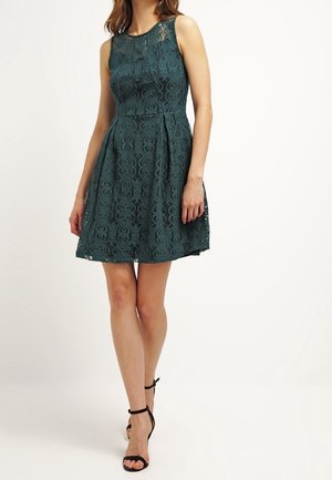 Femme portant une robe sans manches en dentelle vert foncé avec une taille cintrée et des talons noirs à bride à la cheville, debout devant un fond blanc.