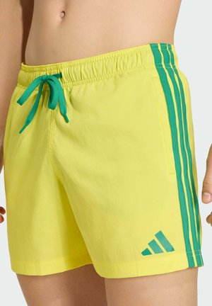 Bañadores amarillos con franjas laterales verdes, cordón verde en la cintura elástica y logo de Adidas verde en la pierna izquierda, usados en la cintura de una persona.