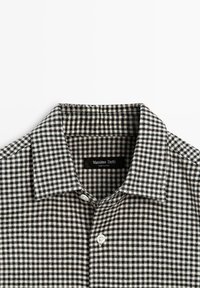 Colletto di camicia a quadri bianco e nero con design appuntito, con etichetta e un bottone bianco nella zona del collo. Tessuto morbido.