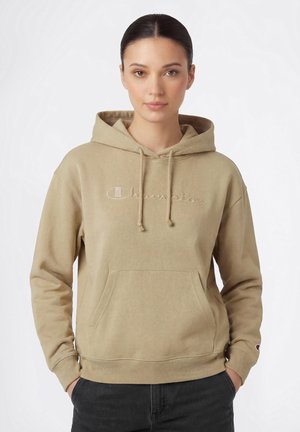 Frau mit dunklen Haaren trägt einen beigen Champion-Hoodie mit Fronttasche und Logo, kombiniert mit schwarzen Hosen, steht vor einem schlichten weißen Hintergrund.