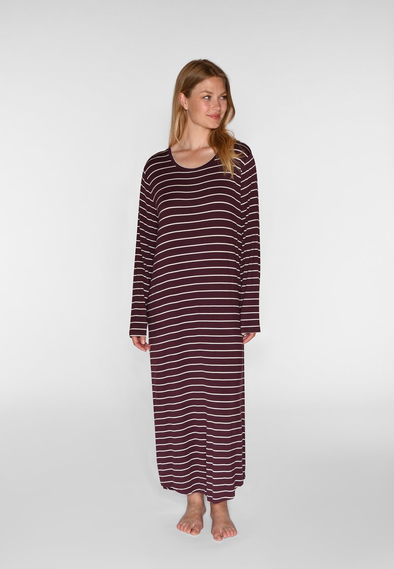 Femme debout pieds nus portant une longue chemise de nuit violet foncé avec des rayures horizontales blanches et des manches longues.