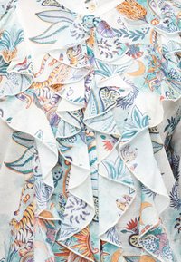 Λευκό πουκάμισο με στρωματοποιημένες βολάν που διαθέτουν πολύχρωμα λουλουδένια, φυλλώδη και τίγρης μοτίβα σε πορτοκαλί, μπλε και πράσινους τόνους.