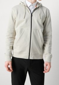 Felpa con cappuccio e zip di colore beige chiaro, dotata di una zip frontale, due tasche anteriori e dettagli della zip nera a contrasto. Tessuto a trama con polsini a costine.