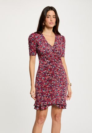 Frau trägt ein eng anliegendes rotes Kleid mit Blumenmuster, kurzen Puffärmeln und einem gerafften Saum, steht vor einem einfarbigen hellen Hintergrund.
