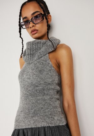 Maglione grigio a maglia con collo alto e design senza maniche. Texture morbida e silhouette aderente. Indossato con occhiali da sole neri oversize.