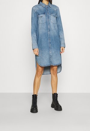 Femme portant une robe en denim bleue à manches longues boutonnée avec des poches poitrine et des bottes de combat noires à lacets, debout sur un fond uni.