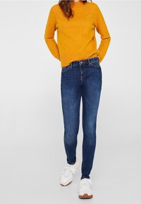 Orange knit sweater with ribbed hem, paired with dark blue skinny jeans. White sneakers with tan accents. Textured fabric details on sweater.