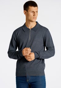 Tummanharmaa polo-paita vetoketjukauluksella, pitkät hihat, ribbaus hihansuissa ja helmassa, valmistettu pehmeästä neulekankaasta. Huomaamaton logoyksityiskohta rinnassa.