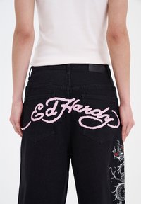 Los jeans de mezclilla negros presentan grandes letras cursivas en color rosa "Ed Hardy" en la parte trasera y un intrincado bordado de dragón en una de las piernas.
