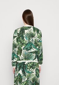 Persoon met lang stijl haar, die een bijpassende groene sweater en rok met tropische bladerenprint draagt, met de rug naar de camera gekeerd tegen een witte achtergrond.