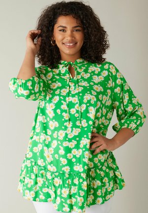 Grüne Blumenbluse mit einem Schlitz-Halsausschnitt, puffigen Ärmeln und einem rüschierten Saum. Ausgestattet mit weißen Gänseblümchen, die auf einem lebhaften Hintergrund gedruckt sind.