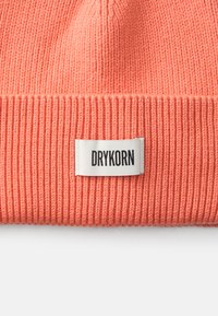 Koralna pletena kapa z rebrasto teksturo in zavihanjem ob robu. Ima pravokotno etiketo z napisom "DRYKORN" v črni barvi.