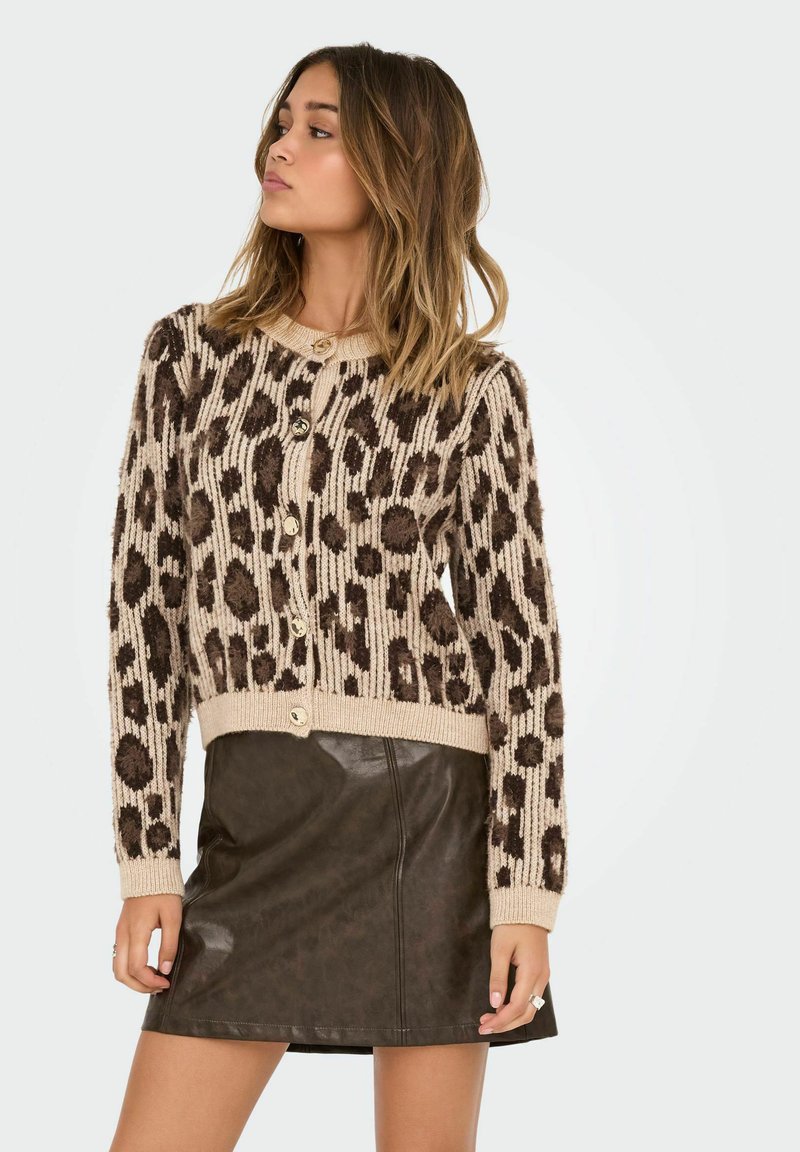 Mlada žena u bež i smeđem kardiganu sa leopard uzorkom na dugmad i tamnosmeđoj kožnoj mini suknji, pozira protiv jednostavne pozadine.