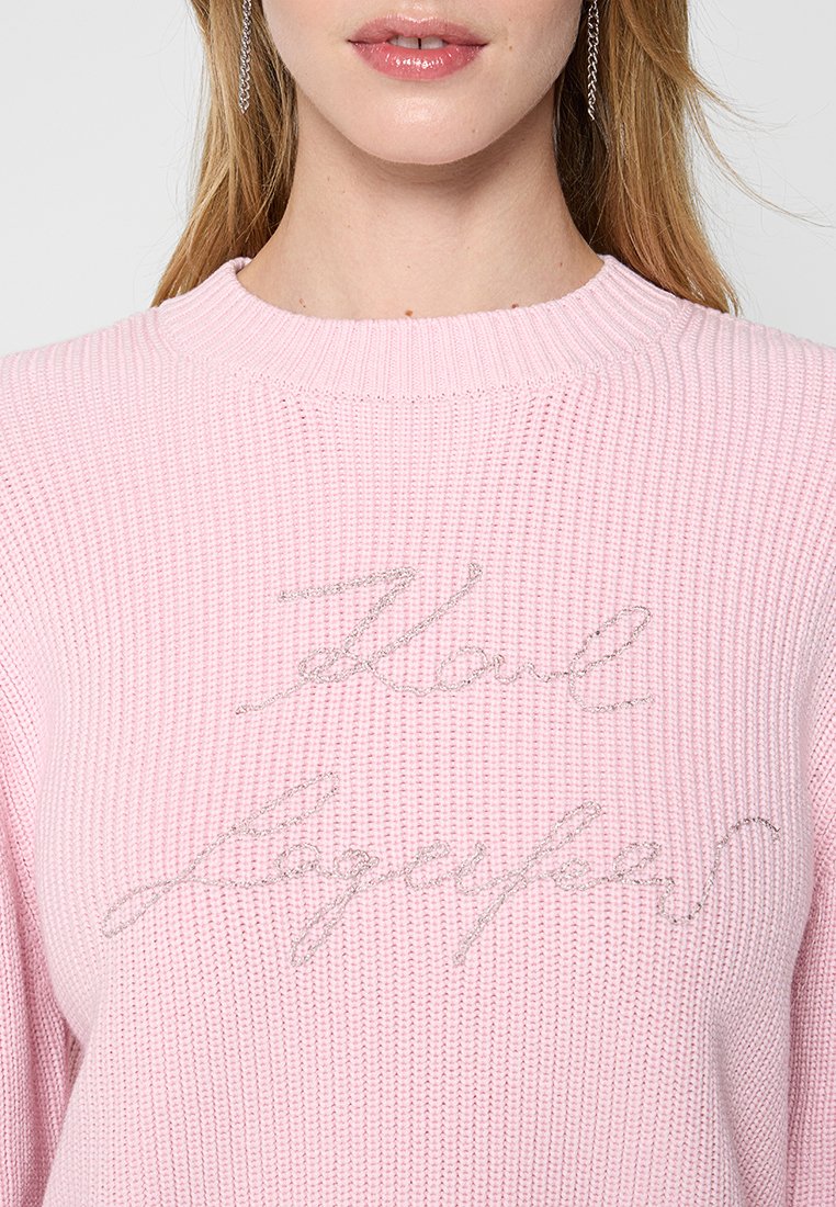 Pull en tricot rose avec un tissage texturé et un col en côtes. Présente un texte brodé argenté sur le devant.