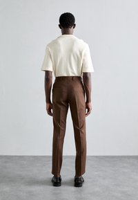 Krämmig texturerad stickad kortärmad topp i kombination med skräddarsydda bruna byxor och svarta skor, sedd bakifrån. Minimalistisk design, inga mönster.