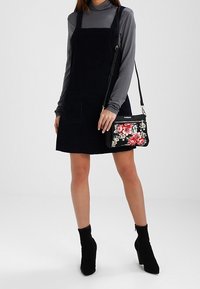 Czarna sztruksowa sukienka ogrodniczka z przednią kieszenią, zestawiona z szarym topem z długim rękawem, czarnymi botkami do kostki oraz torebką w kwiatowy wzór.