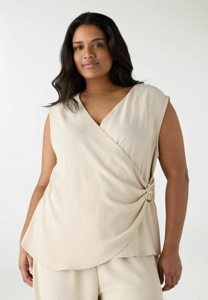 Donna con capelli scuri che indossa un top beige senza maniche con cintura dorata e pantaloni beige abbinati, in piedi su sfondo bianco.