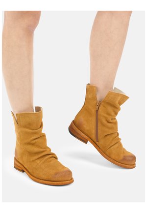 Lichtbruine suède enkellaarsjes met een slappe vormgeving en een ritssluiting aan de zijkant, gedragen op blote benen tegen een witte achtergrond.