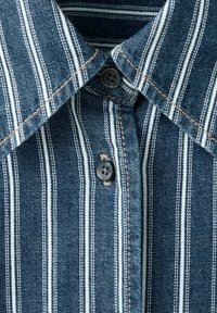 Chemise en denim bleu avec des rayures verticales blanches, col visible et deux boutons noirs fermés sur la patte de boutonnage avant.
