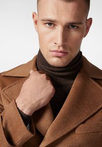 Brązowy wełniany płaszcz z wyprofilowanym kołnierzem, założony na ciemnobrązowy golf. Widoczna dłoń trzymająca klapę płaszcza, ukazująca fakturę i detale krawieckie.