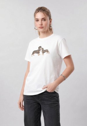 Νέα γυναίκα με αλογοουρά που φοράει λευκό μπλουζάκι με δύο εικόνες από σκύλους ντάχσχουντ, μαύρα τζιν και ασημένια σκουλαρίκια κρίκους και βραχιόλι.