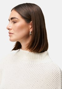 Gebreid trui in zacht crème met ribbeltextuur en een hoge kraag; model draagt zilveren ringoorbellen, die de glanzende, rechte afwerking van het haar accentueren.