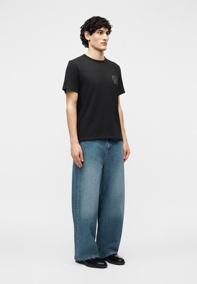 Νέος άνδρας που φοράει μαύρο μπλουζάκι με μικρό λευκό κυκλικό λογότυπο και ευρύχωρα μπλε τζιν, στέκεται μπροστά σε απλό φόντο.