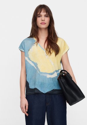 Frau mit langen braunen Haaren, die ein Oberteil mit blau-gelbem abstraktem Muster und dunkle Jeans trägt, in ihrer linken Hand eine schwarze Ledertasche haltend.