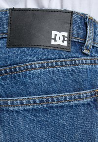 Džínsy z denimu s modrým praním, viditeľným šitím a čiernou koženou nášivkou s bielym logom "DC" na páske.