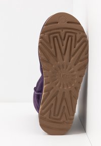Semelle d'une botte UGG violette montrant un motif de bande de roulement détaillé et le logo de la marque sur un fond blanc.