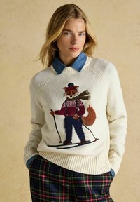 Balts adīts džemperis ar izšūtu lapsu, kas slēpo, ar bordo džempera rakstu, apvienots ar džinsa apkakli un raibām biksēm.