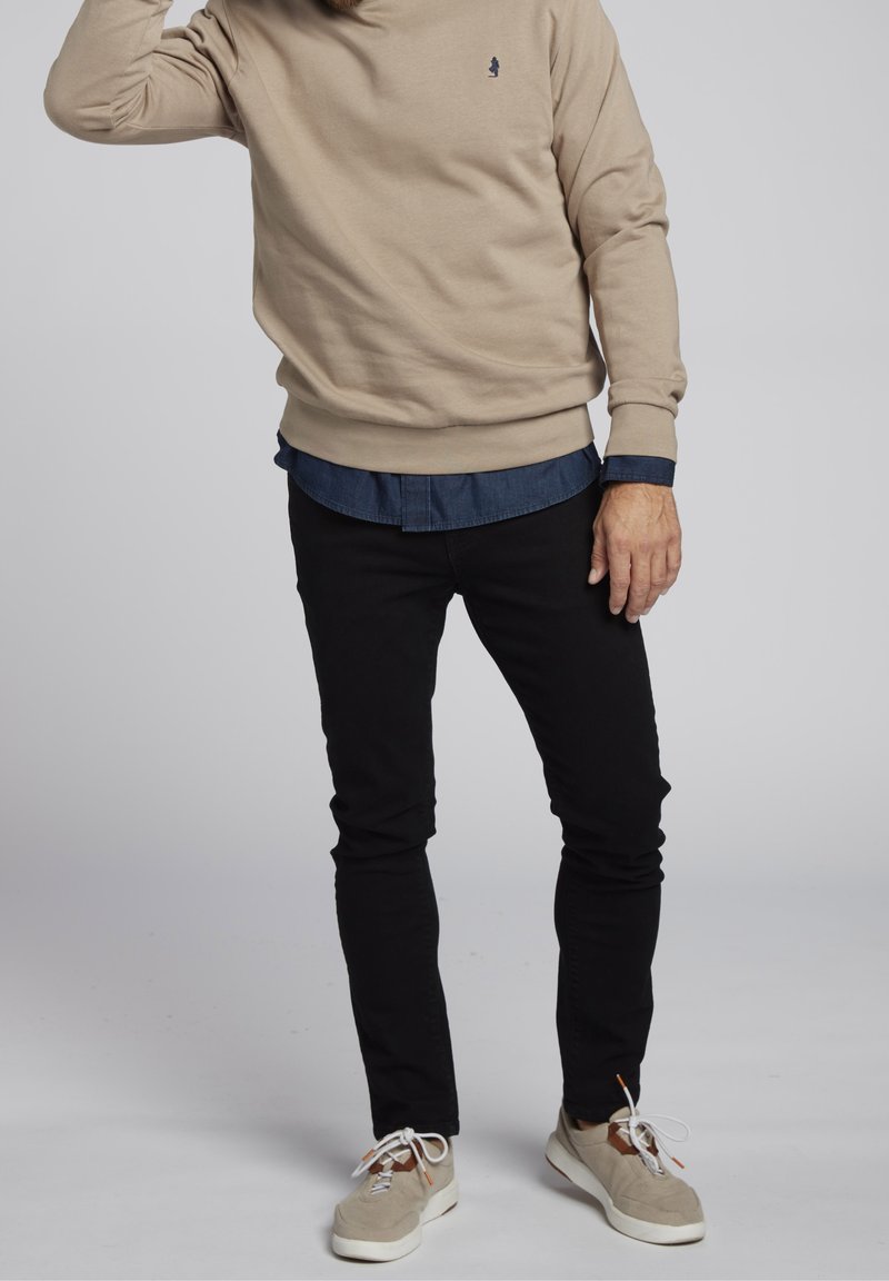 Homme portant un sweat-shirt beige sur une chemise bleue, un pantalon noir et des baskets beiges, debout sur un fond uni gris clair.