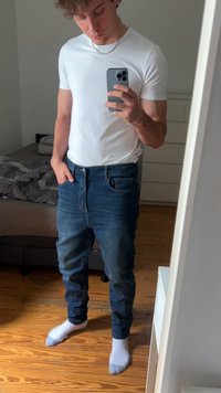 Mlad moški v beli majici in modrih kavbojkah dela selfie v ogledalu v spalnici z lesenim podom in neurejeno posteljo.